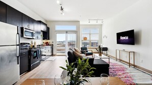 Un apartamento moderno con cocina, comedor y sala de estar. Cuenta con una televisión de pantalla plana, un ventanal grande y un balcón.