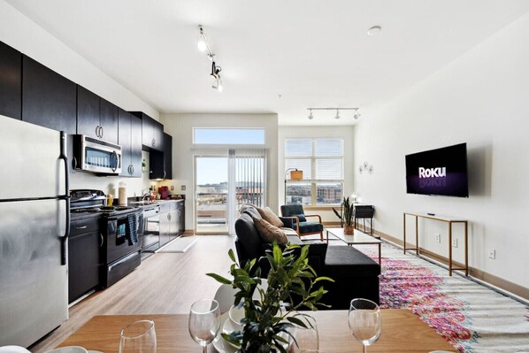 A modern apartment with a kitchen, dining area, and living room. It features a flat-screen TV, a large window, and a balcony. - 1Br Apt in Deep Ellum with King bed, Pool & Outdoor Space (Dallas)