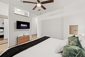 A modern hotel room with a bed, a TV mounted on the wall, a wooden dresser, and a ceiling fan. - Tasteful Apt in Central Dallas with Parking and Pool (Dallas)