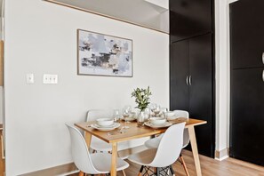 A dining area with a wooden table set for four, white chairs, a vase with greenery, and a framed abstract painting on the wall. - Tasteful Apt in Central Dallas with Parking and Pool (Dallas)