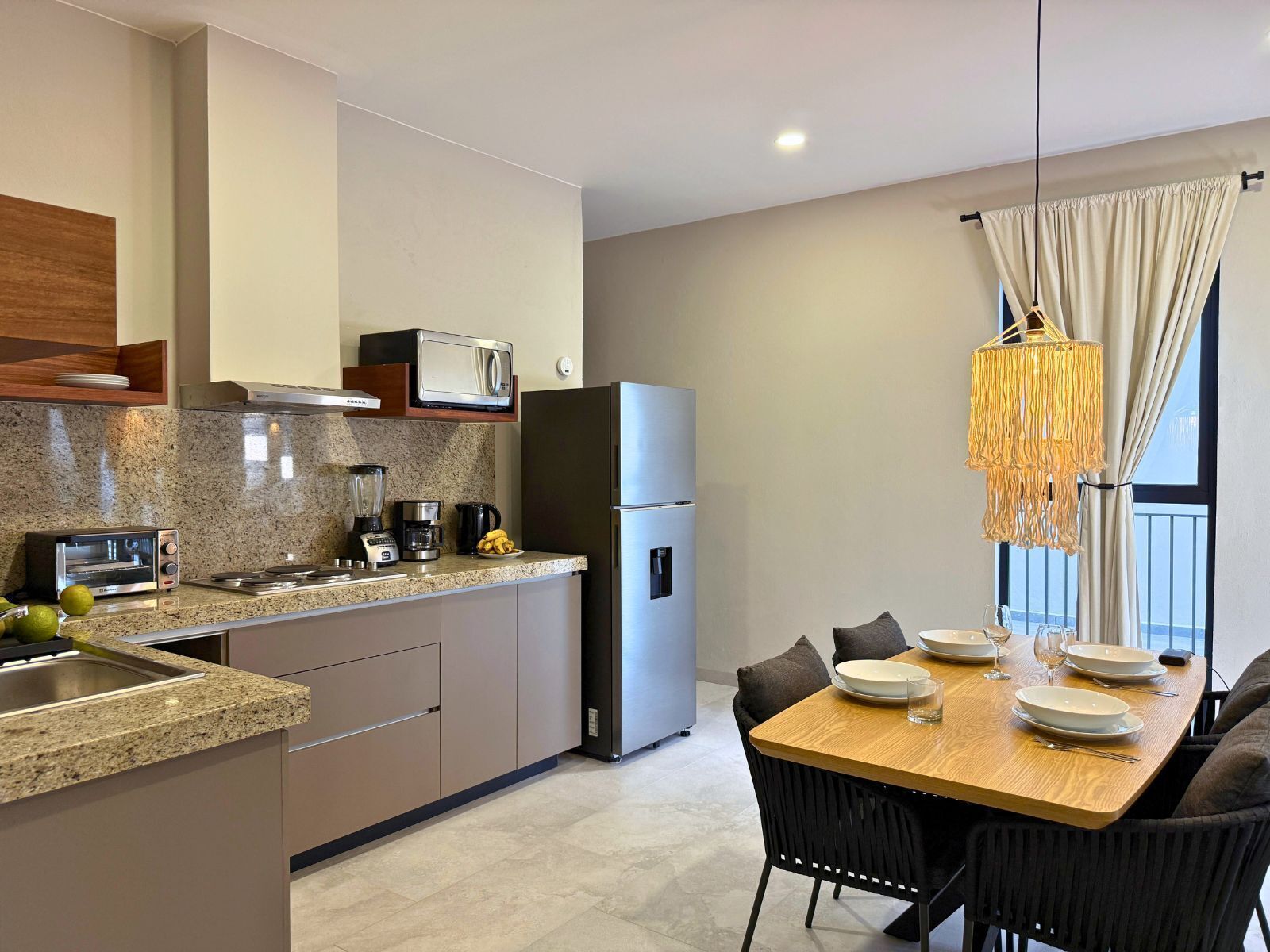 A modern kitchen with granite countertops, a built-in microwave, a stainless steel refrigerator, and a dining area with a wooden table and black chairs.