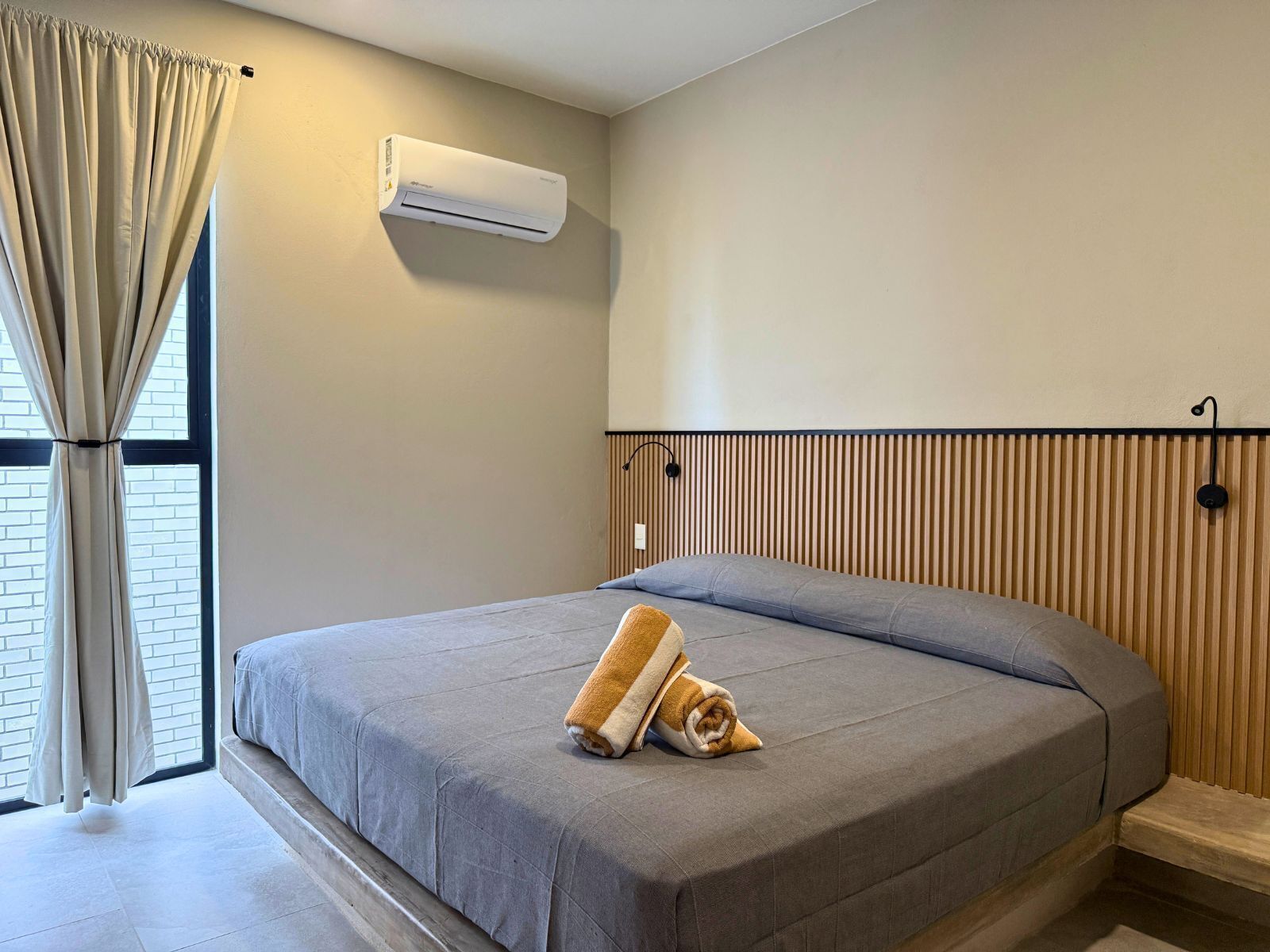 A modern hotel room with a bed, a window with curtains, a wall-mounted air conditioner, and a wooden panel wall.