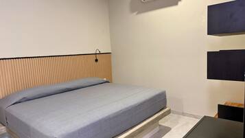 Un lit simple avec un matelas gris dans une pièce dont le mur est blanc, avec une tête de lit en lattes de bois et une table noire.