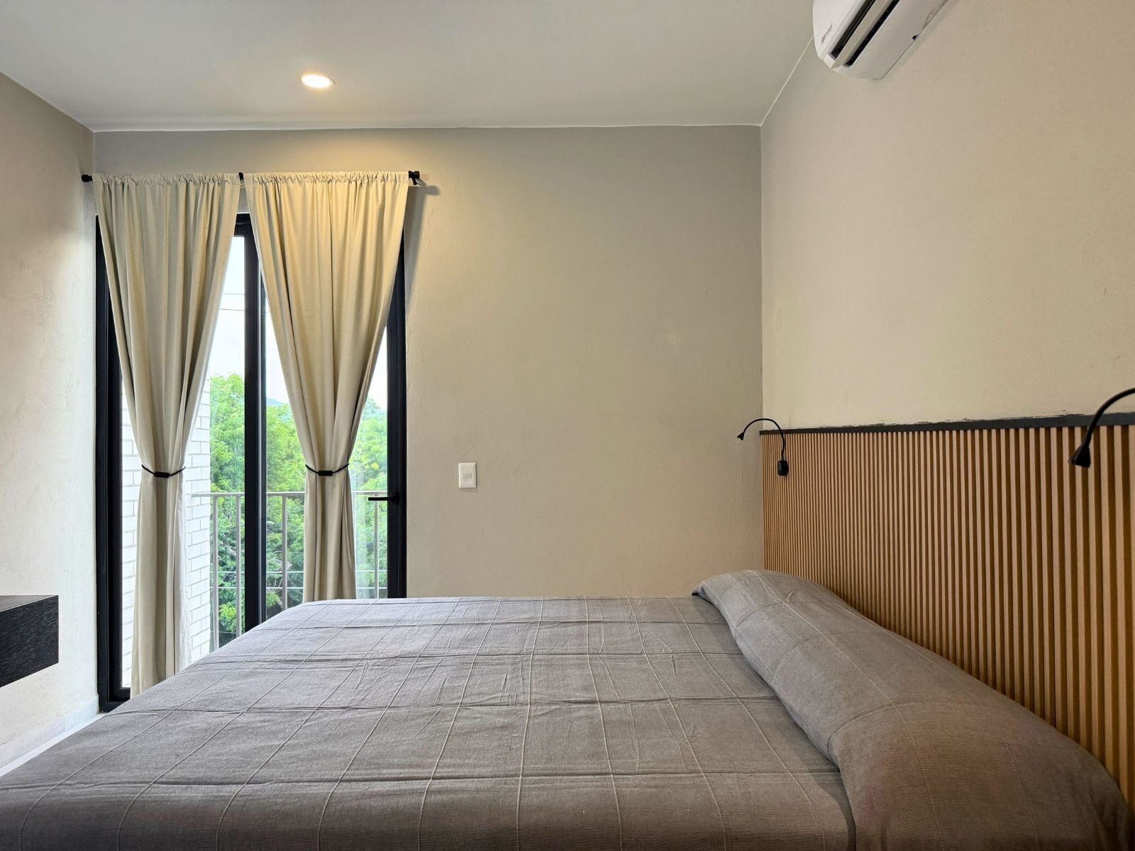 Un dormitorio con una cama grande, cortinas color beige, un radiador y vistas al exterior con vegetación.