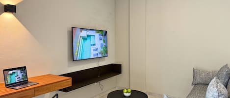 Un salon moderne avec un téléviseur à écran plat fixé au mur, un bureau en bois avec un ordinateur portable, une table basse ronde noire sur laquelle se trouve un saladier de fruits, et un canapé gris à motifs.