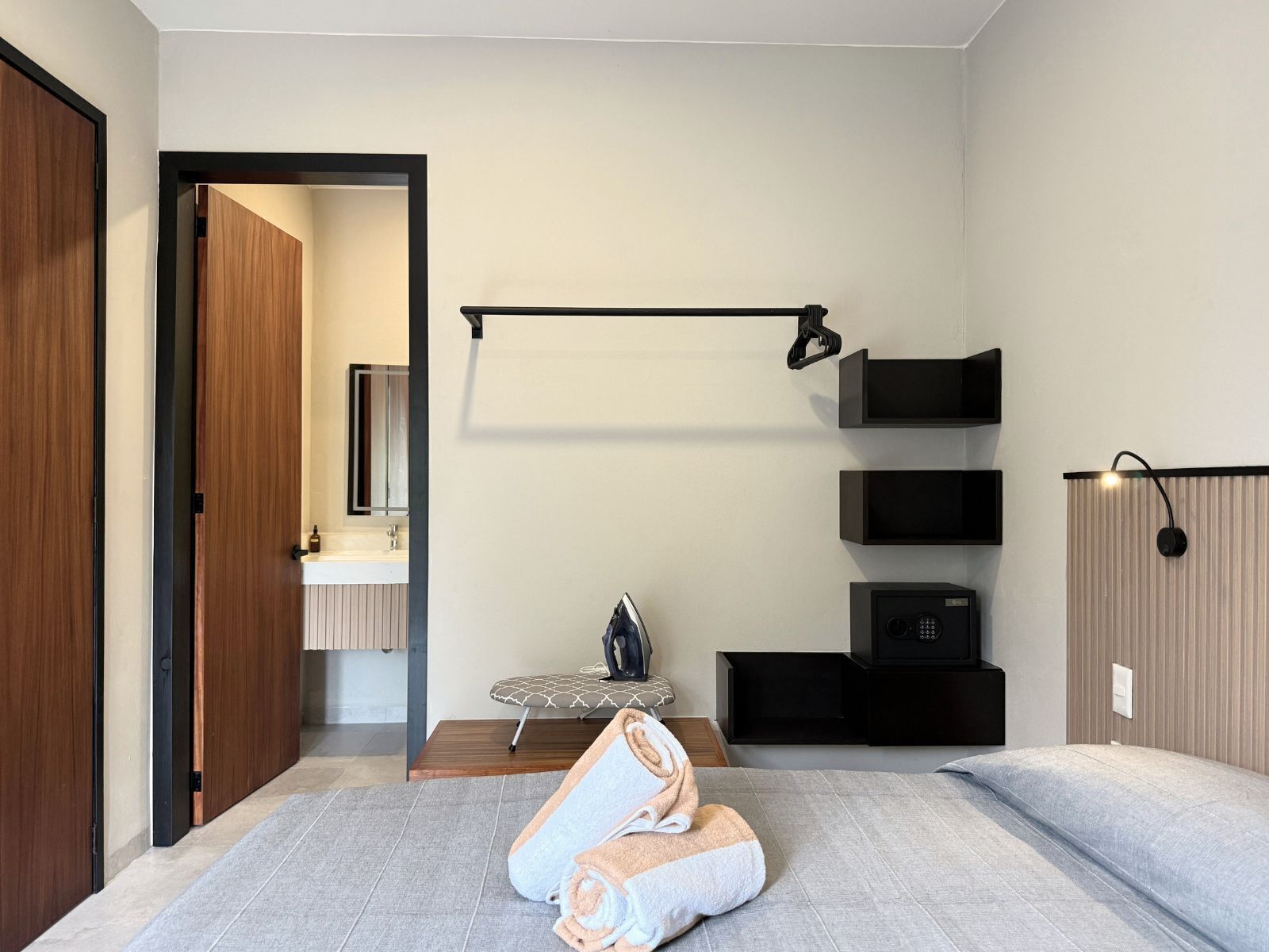 A modern hotel room with a bed, a small seating area, a vanity with a mirror, and a wall-mounted shelf unit.