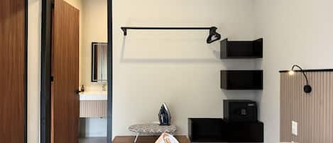 A modern hotel room with a bed, a small seating area, a vanity with a mirror, and a wall-mounted shelf unit.