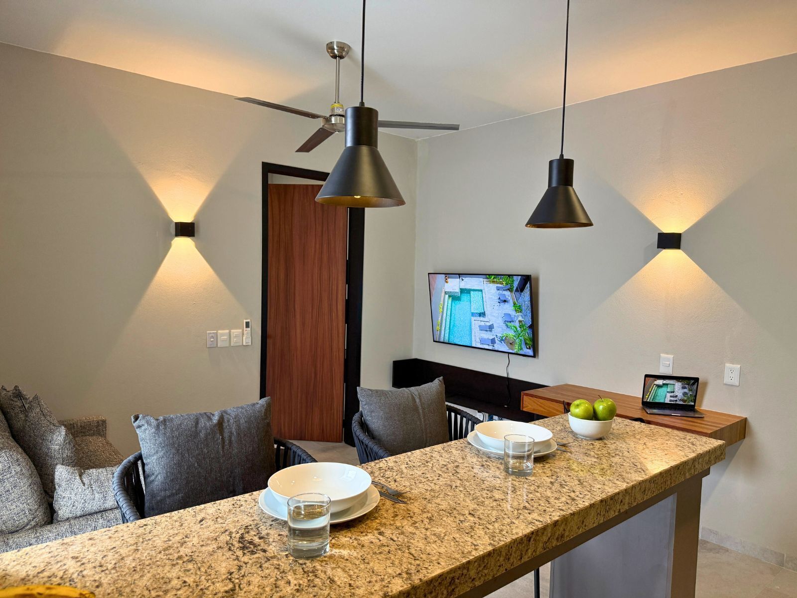 Eine moderne Küche mit Granit-Arbeitsplatten, ein Essbereich mit grau gepolsterten Stühlen und ein an der Wand montierter Fernseher, der einen Blick auf einen Pool zeigt.