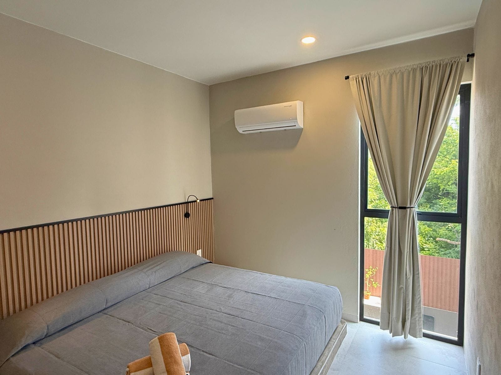 A bedroom with a bed, a window with curtains, a wall-mounted air conditioner, and a wooden slat headboard.
