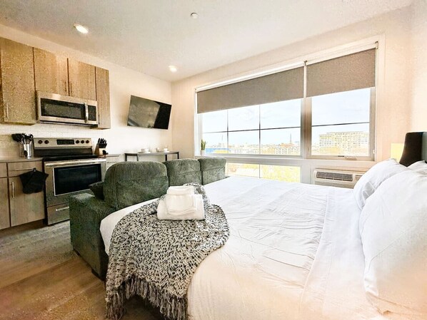 Une chambre d’hôtel moderne dotée d’une kitchenette, d’un lit avec du linge de maison blanc, d’un fauteuil vert et d’une grande fenêtre donnant sur la ville.