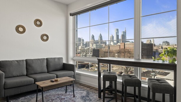 Un salon moderne avec un canapé gris, une table basse en bois et une vue sur la ville grâce à de grandes fenêtres.