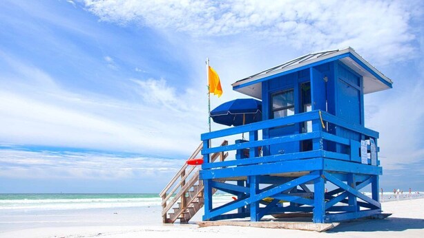 Una torre azul de salvavidas con una bandera, escaleras y una playa al fondo.