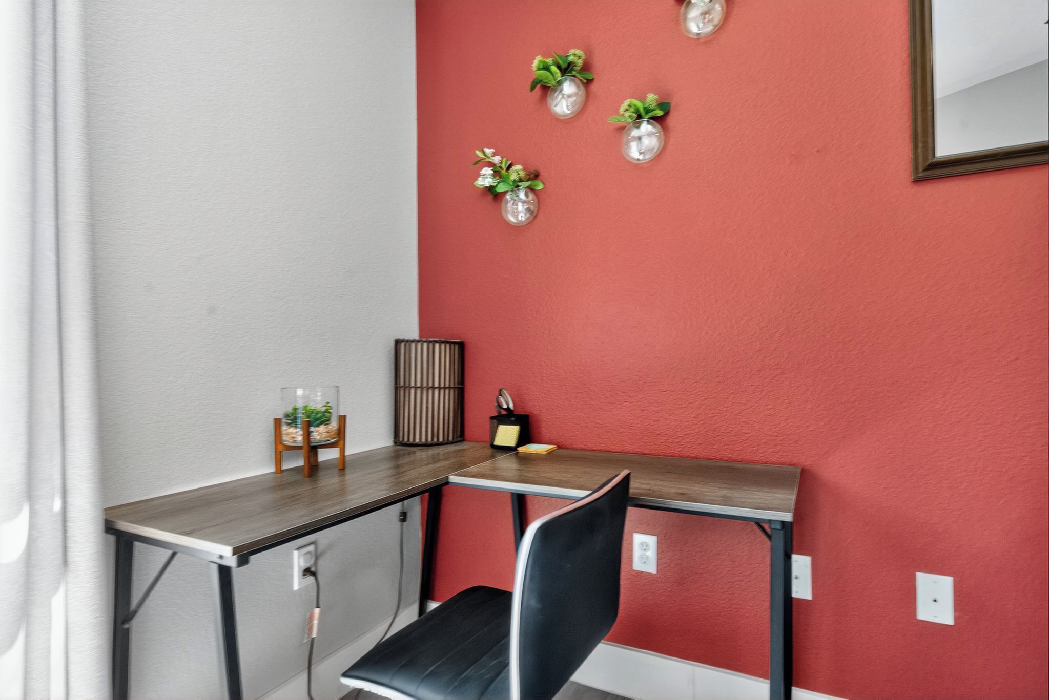 Een hoekbureau met een zwarte stoel, een klein plantje op een voetstuk en een spiegel aan de muur.