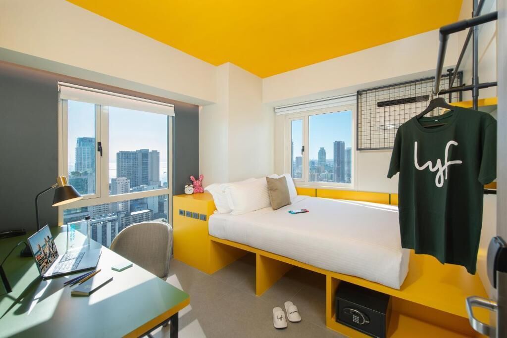 Une chambre d’hôtel moderne équipée d’un lit, d’un bureau avec un ordinateur portable et d’une fenêtre donnant sur la ville.