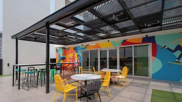 An outdoor seating area with yellow chairs and tables, a colorful mural, and a glass door entrance.