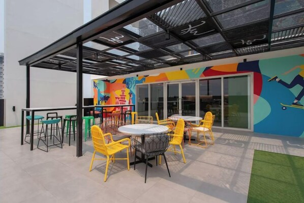 Un espace extérieur aménagé avec des chaises et des tables jaunes, une fresque murale colorée et une entrée avec une porte en verre.