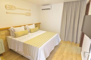 A hotel room with a bed, pillows, a nightstand, an air conditioning unit, and a curtain. - Pousada Topazio (Cairu)
