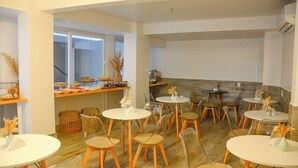 A dining area with round tables and chairs, a kitchen area with a counter and various items, and a view of the outdoors through large windows. - Pousada Topazio (Cairu)