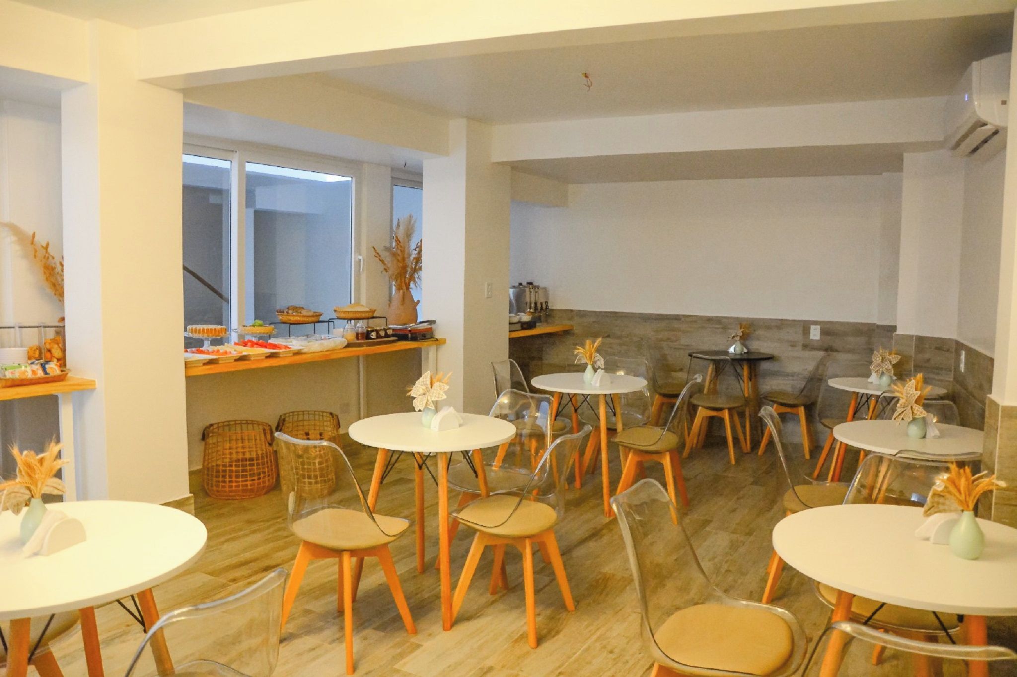 Un comedor con mesas y sillas redondas, una zona de cocina con mostrador y varios objetos, y vista al exterior a través de amplios ventanales.