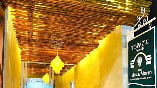 A corridor with a wooden ceiling, yellow walls, and a sign for Topazio Pousada. - Pousada Topazio (Cairu)
