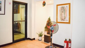 A hotel lobby with a glass door, a standing fan, a small Christmas tree, and a framed religious painting.