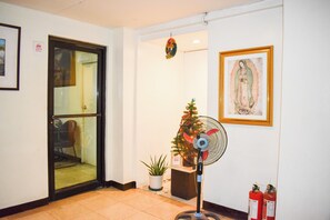 A hotel lobby with a glass door, a standing fan, a small Christmas tree, and a framed religious painting.