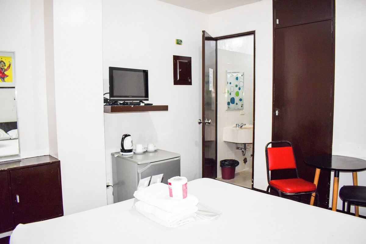 A hotel room with a bed, a desk with a television, a small kitchenette, a bathroom, and a seating area with a table and chairs.