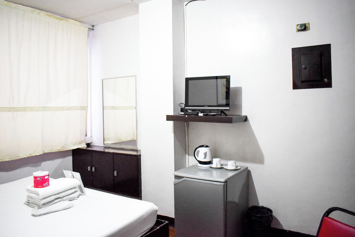 A hotel room with a bed, a desk with a computer, a TV mounted on the wall, a small refrigerator, and a red chair.