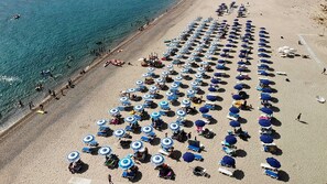 Vista aérea de una playa con filas de tumbonas y sombrillas, cielo despejado y el océano.