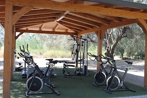 Un espacio exterior techado para gimnasio con bicicletas estáticas, una banca y equipo de levantamiento de pesas.