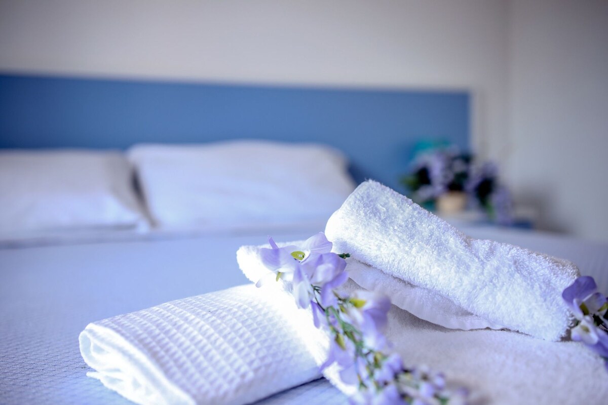 Una cama bien hecha con sábanas blancas y una toalla doblada, acompañada de flores moradas.