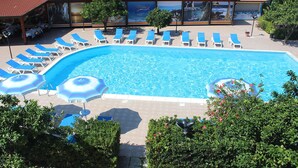 Piscina con sillones de descanso y sombrillas, rodeada de follaje y un edificio de techo rojo.