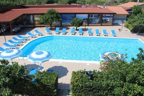 Piscina con sillones de descanso y sombrillas, rodeada de follaje y un edificio de techo rojo.