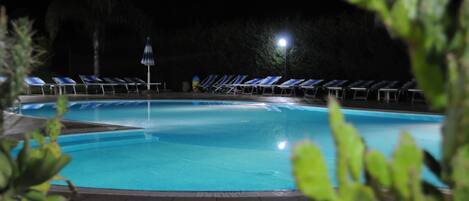 A swimming pool with blue lounge chairs and a light post at night.