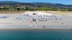 Vista aérea de una playa con numerosas sombrillas y tumbonas, un barco en el agua y montañas al fondo.