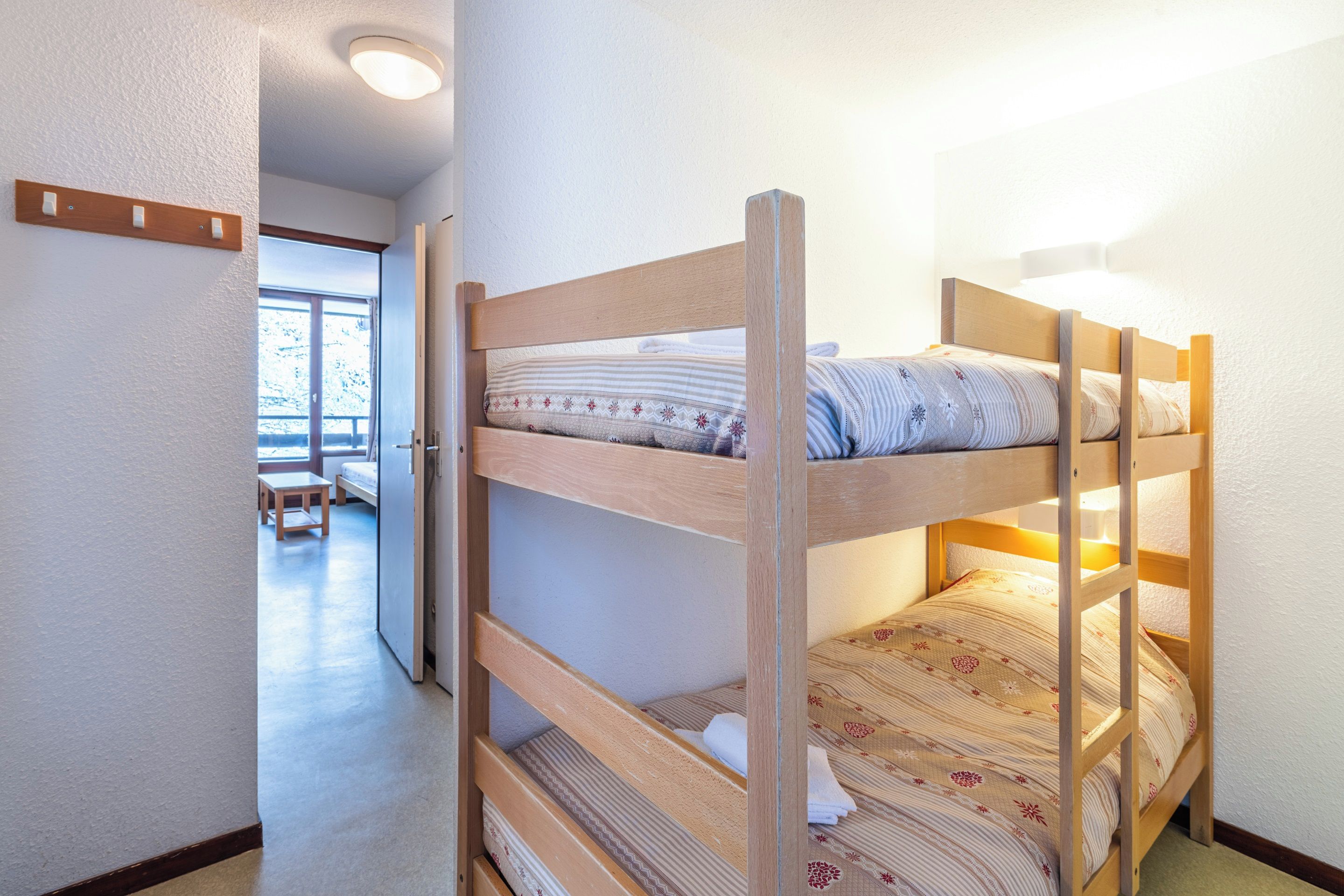 Una cama litera con estructura de madera, una colcha estampada y una lámpara de noche. En la habitación contigua hay una mesita y un banco.