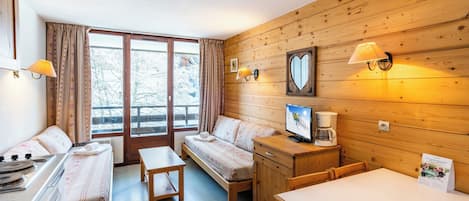 A cozy cabin-style room with a bed, a desk, a chair, a table, a stove, and a window with curtains.