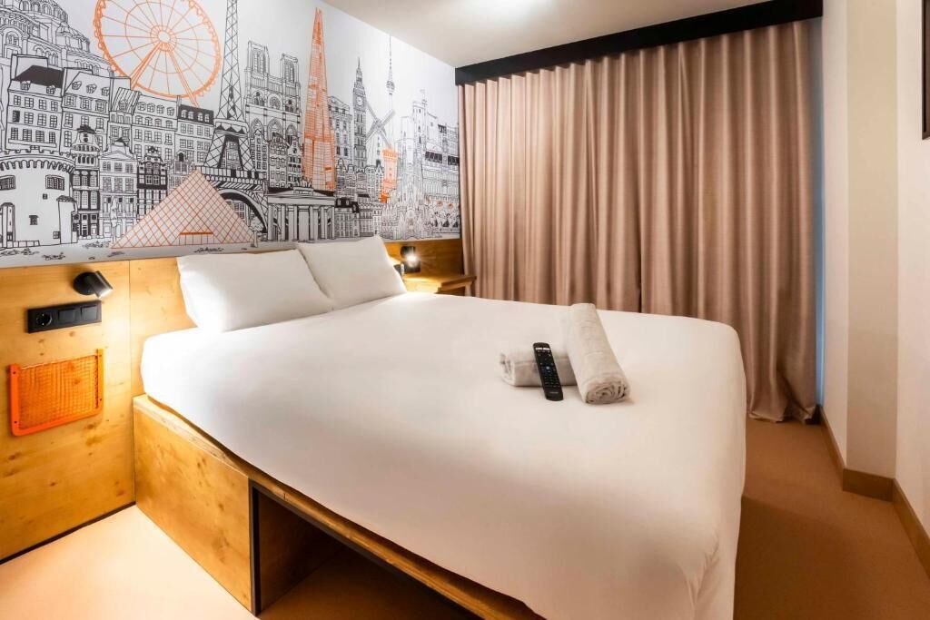 A hotel room with a large bed, a headboard with a cityscape mural, a wooden nightstand, a lamp, and a window with curtains.