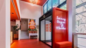 A modern interior with a red-orange floor, a neon sign reading "I am in my Orange era Barcelona," and a staircase leading to a second floor.