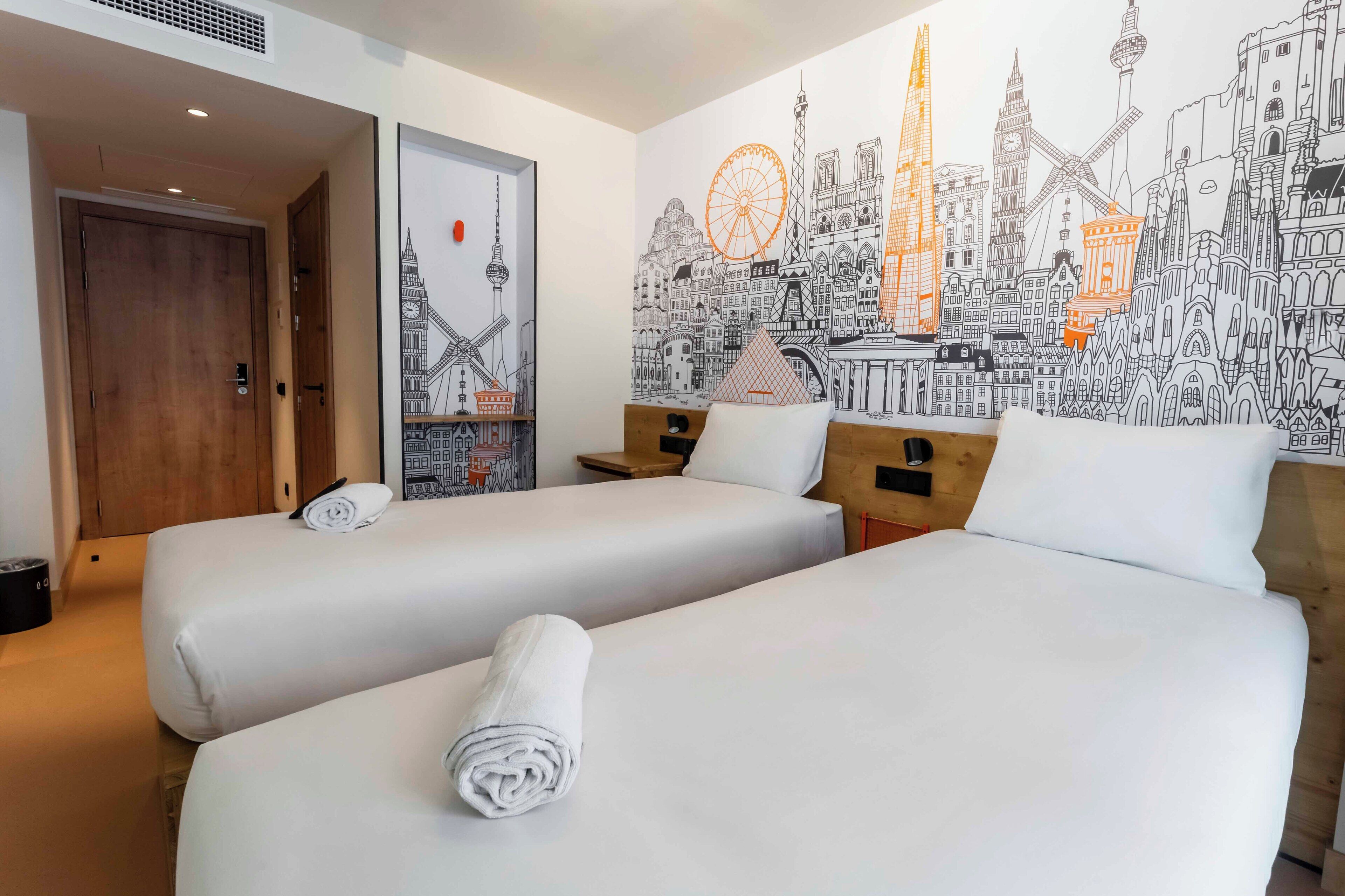 A hotel room with two beds, a cityscape mural, and a wooden door.