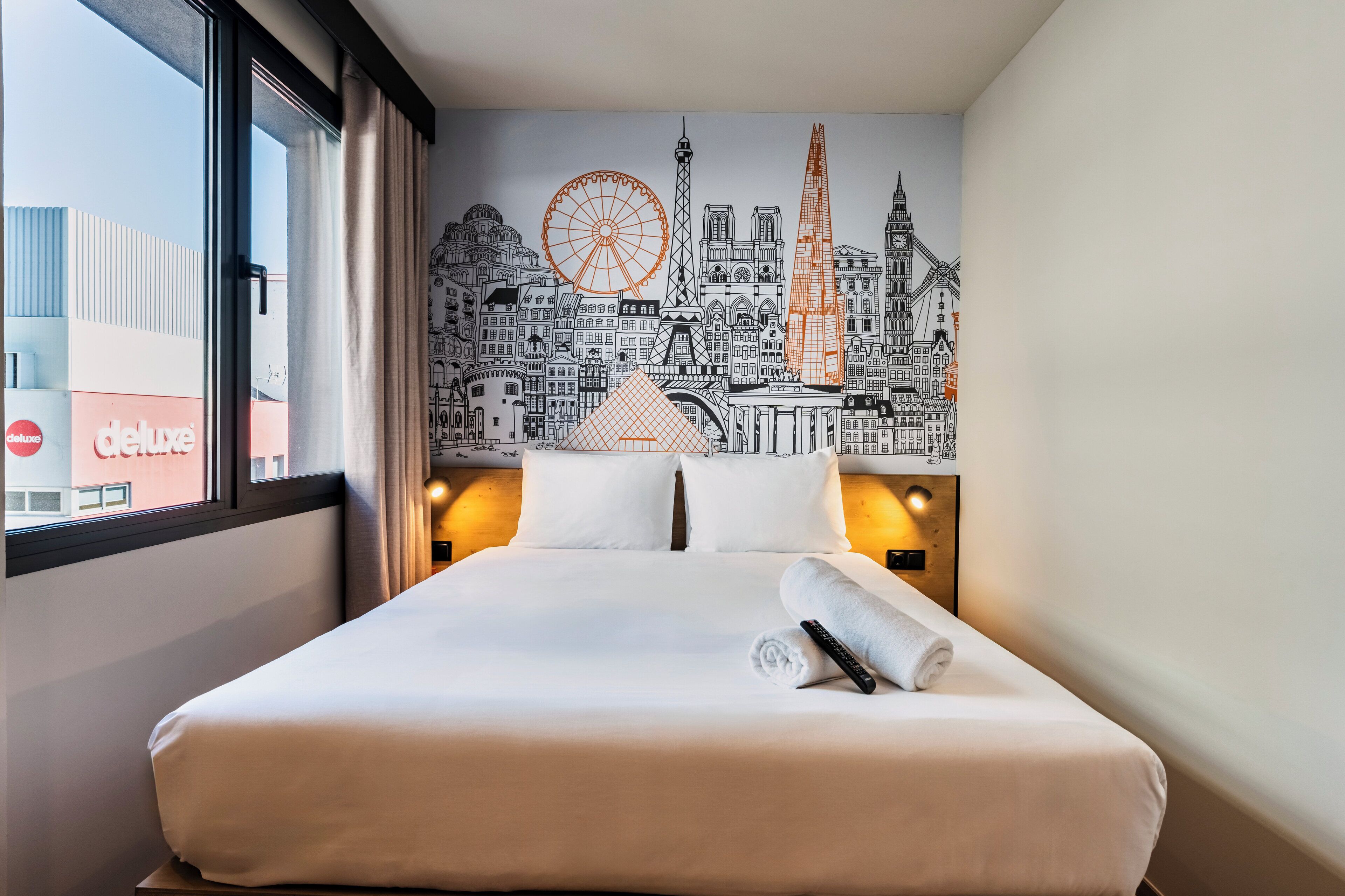 A hotel room with a large bed, a cityscape mural on the wall, and a window with a view of buildings.