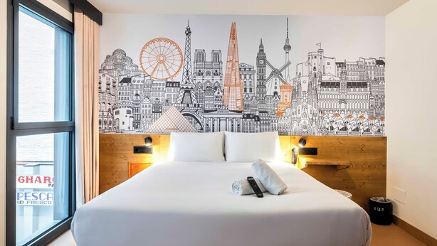 A hotel room with a large bed, a cityscape mural on the wall, and a window with curtains.