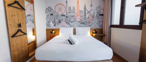 A hotel room with a large bed, wooden headboard, a cityscape mural, a wooden wardrobe, and a window with curtains.