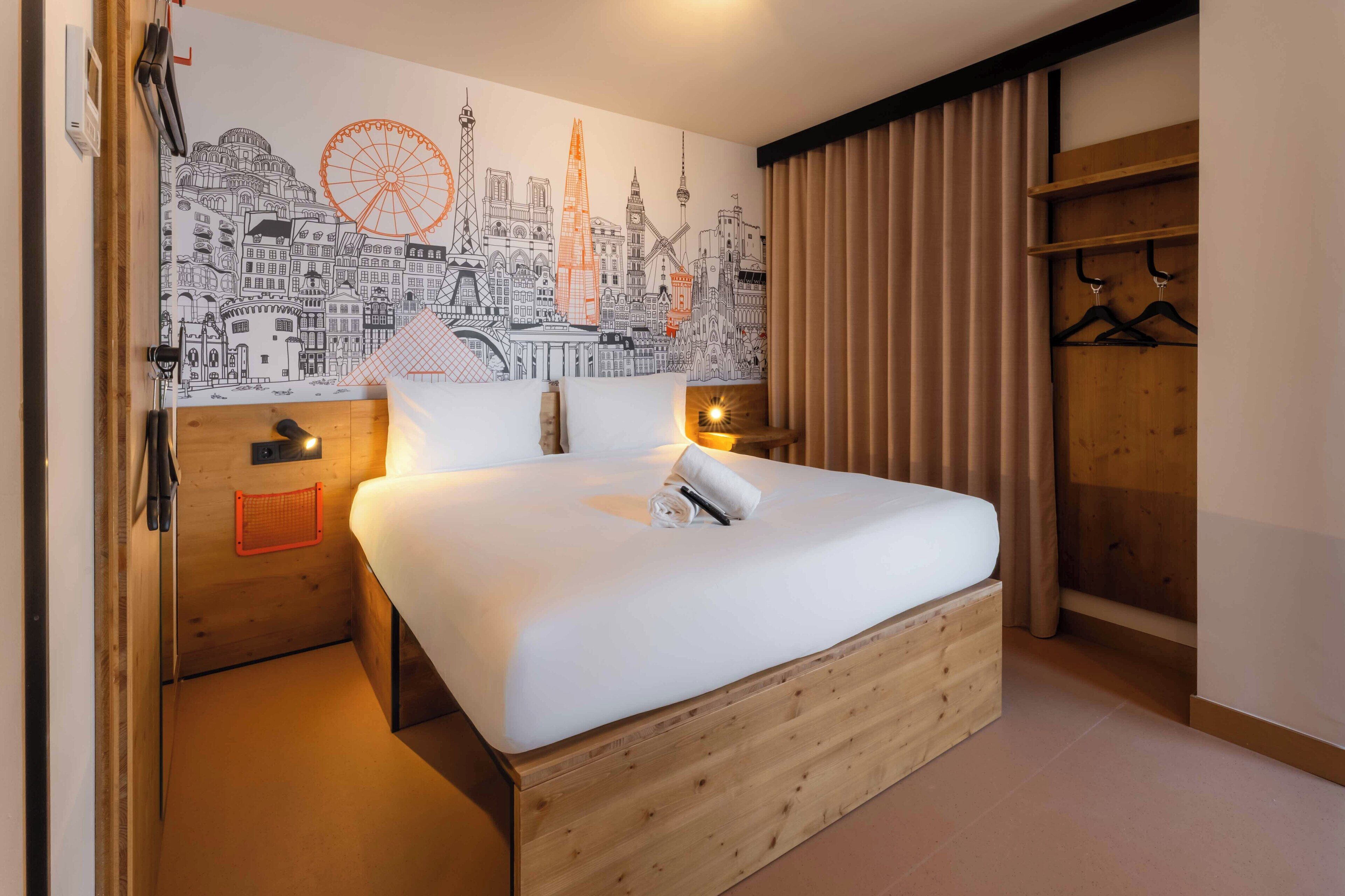A hotel room with a large bed, wooden headboard, a cityscape mural on the wall, a wooden wardrobe, and a small shelf with a towel.