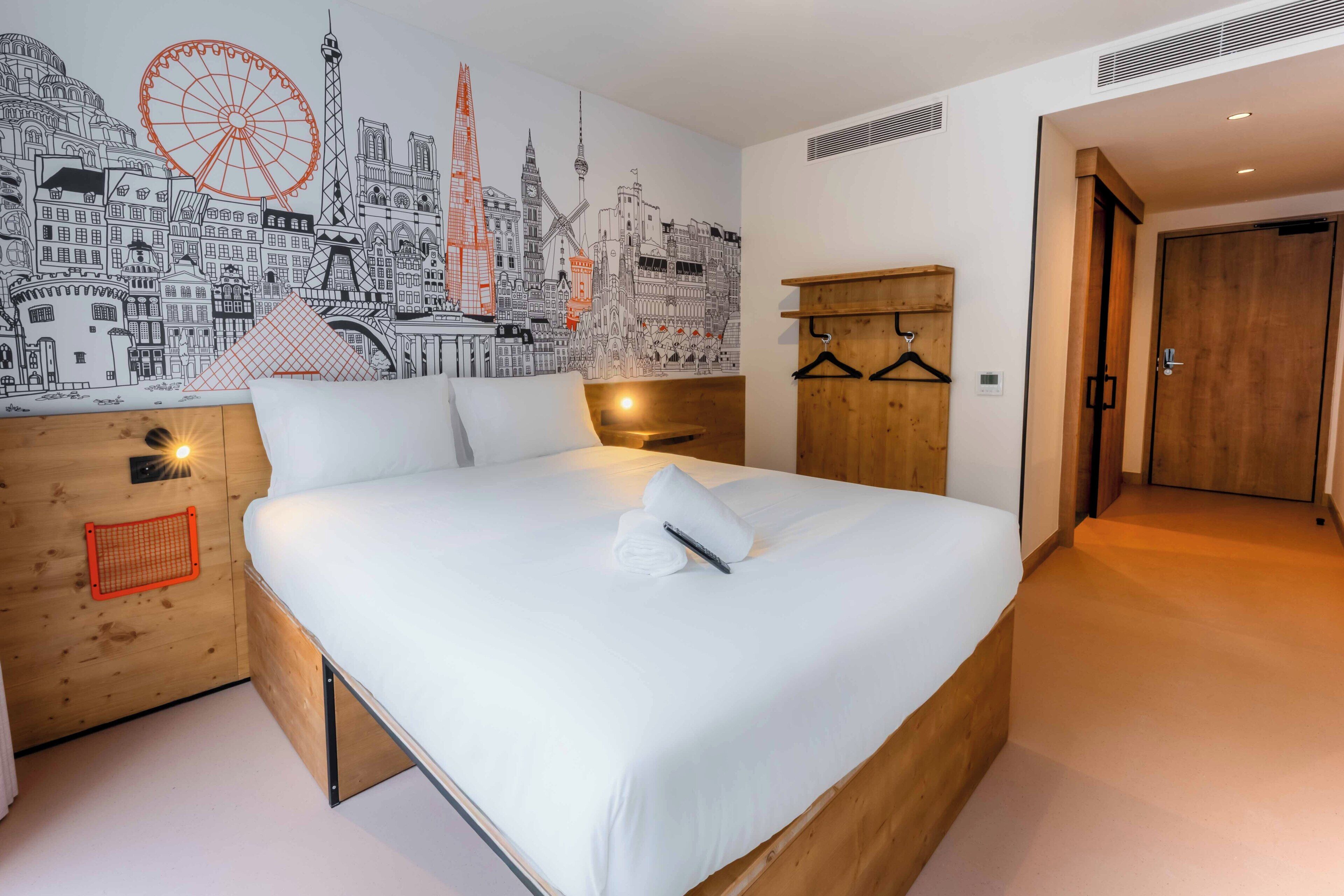 A modern hotel room with a large bed, a cityscape mural, a wooden headboard, a wooden wardrobe, and a wooden bench.