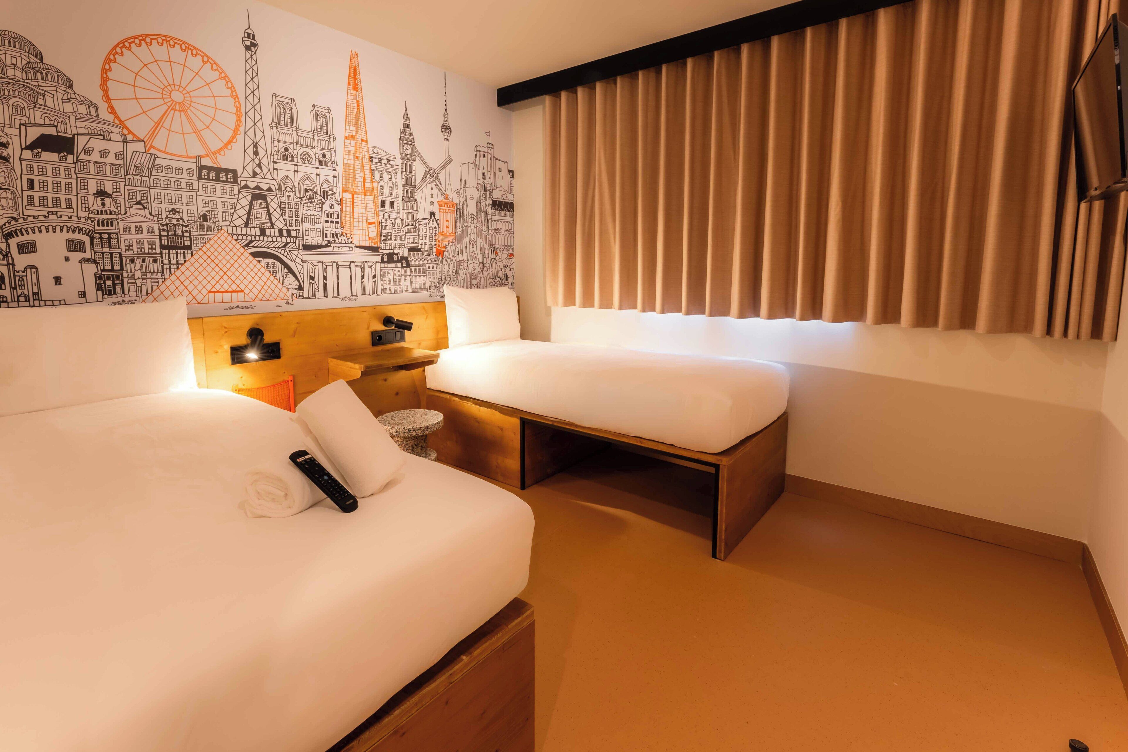 A hotel room with a cityscape mural, a bed, a bedside table with a lamp, and a window with curtains.