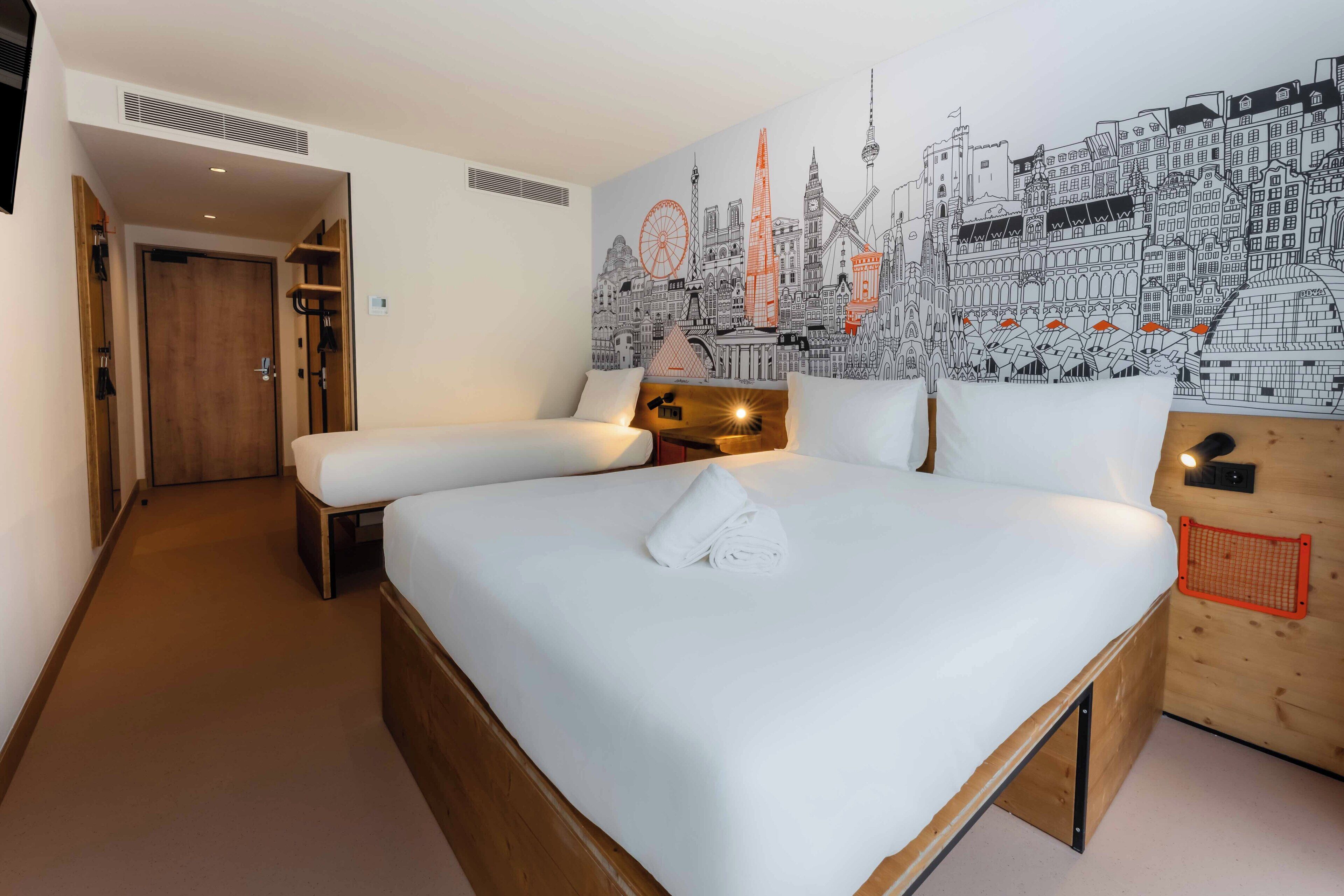 Habitación de hotel con una cama grande, dos camas más pequeñas, un mural con vista a la ciudad y un cabecero de madera.