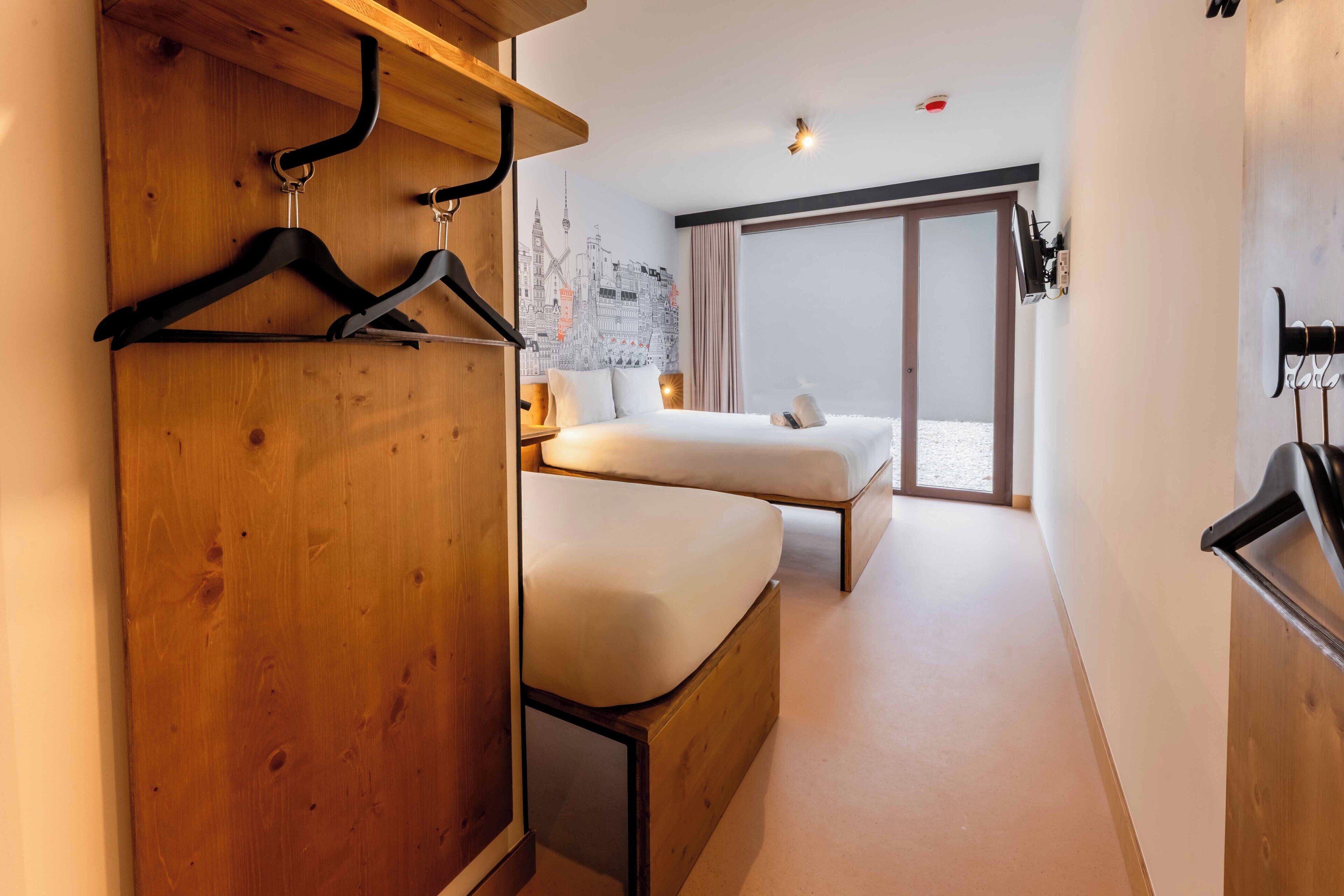 A hotel room with a wooden wardrobe, two beds, a cityscape mural, and a sliding glass door to the outside.