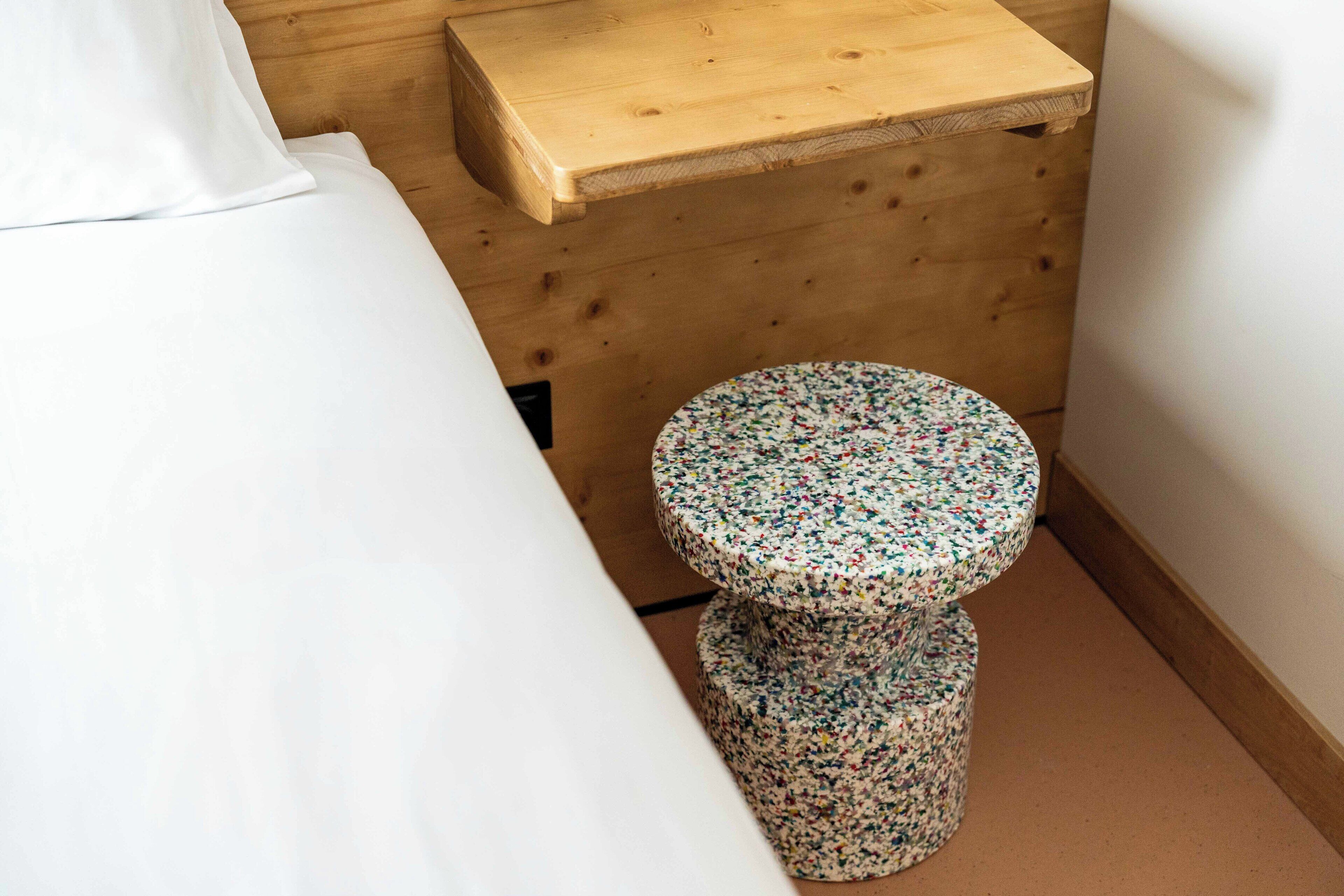 A small, round stool with a floral pattern, a wooden bench, and a white bed in a room with wooden walls.