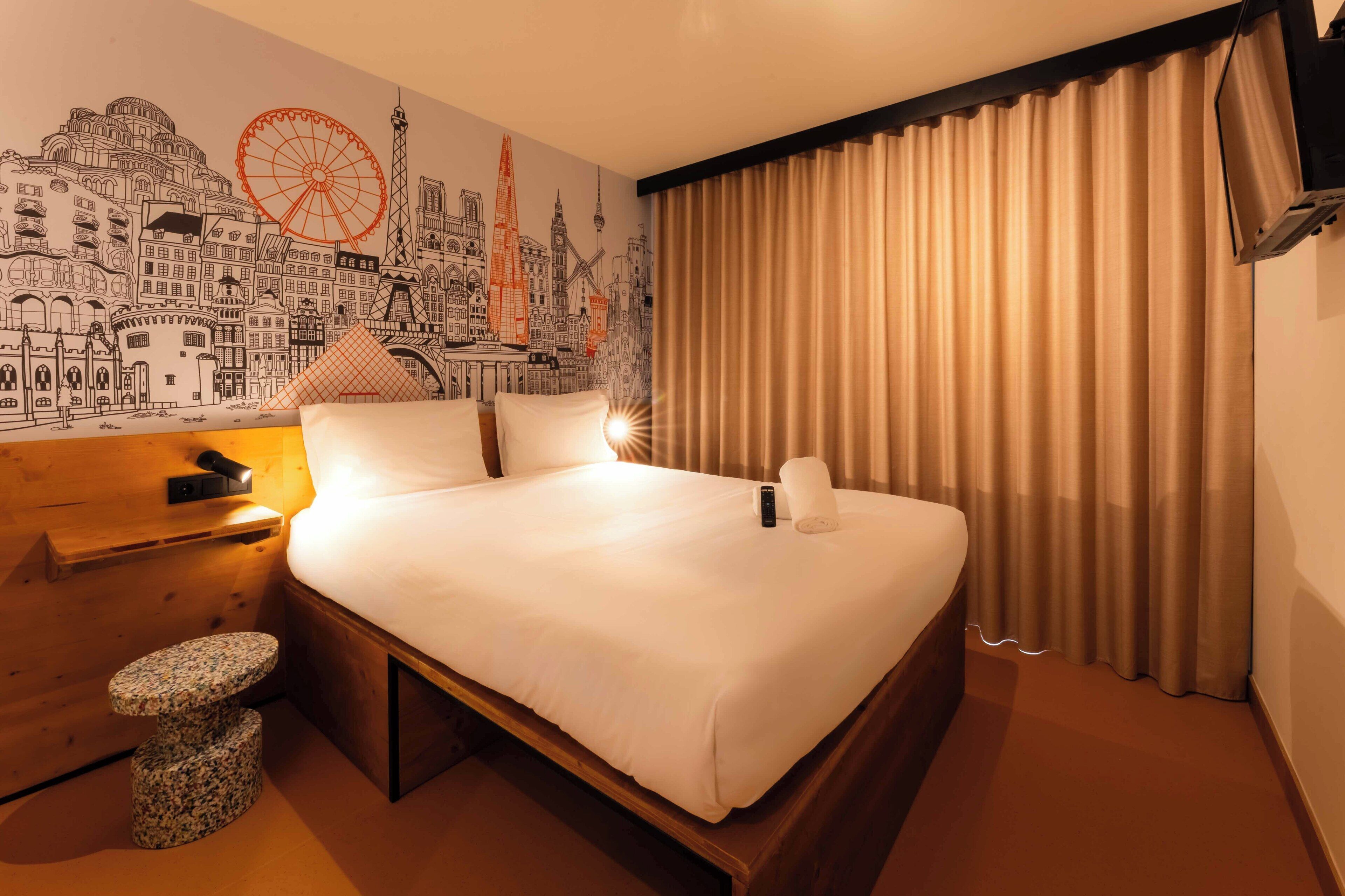 Une chambre d’hôtel avec un grand lit, une table de chevet en bois, un petit tabouret rond, une fresque murale représentant un paysage urbain et une grande fenêtre avec des rideaux.